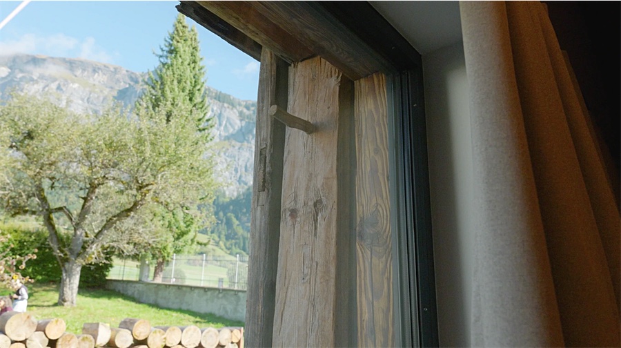 Alter Balken des Stalls neben dem neuen Fensterprofil, dahinter Obstbaum und Berge