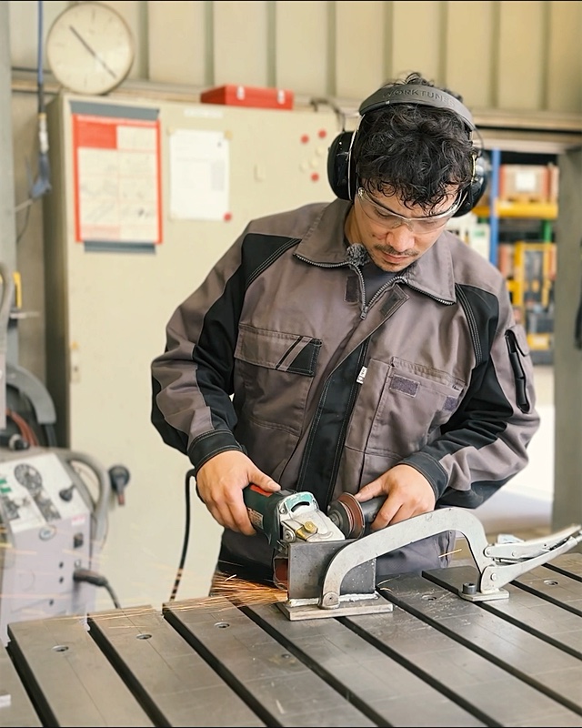 Metallbauer mit Gehörschutz schleift ein Bauteil in der Werkstatt von HIRT kinetics